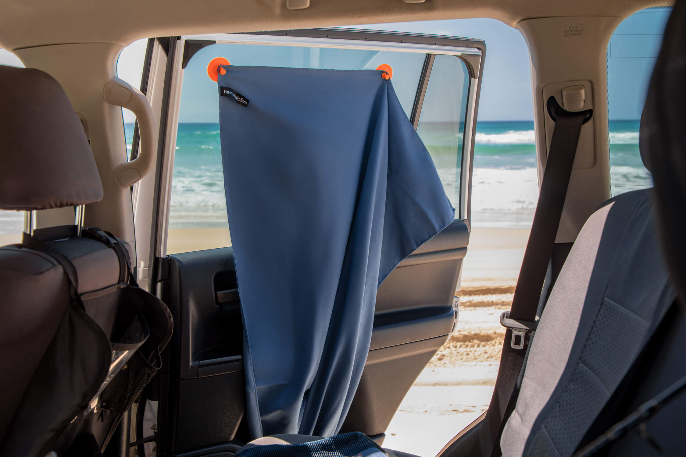 Car interior with a blue Fortunate Sun Hidey towel hung with suction cups over the window acting as a privacy screen, with the beach visible through the window.