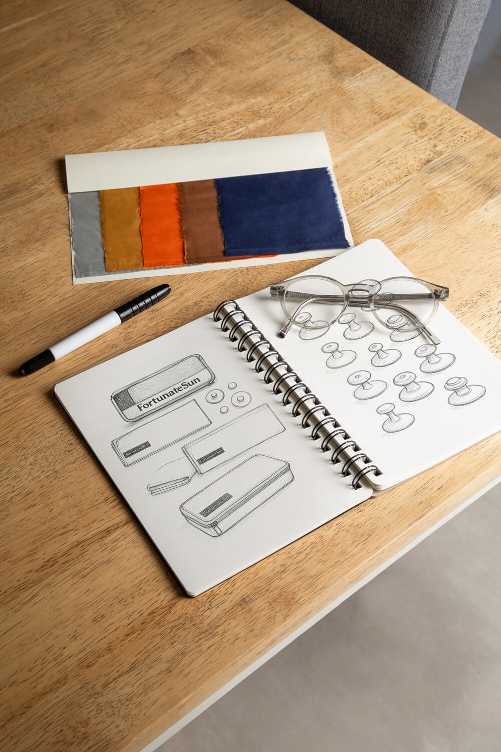 Fabric swatches on a table next to a book of product sketches of the Hidey towel by Fortunate Sun with glasses sitting on the top.